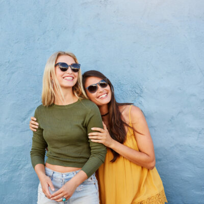 Portrait of two young women standing together over blue background. Stylish young female models looking away and smiling.